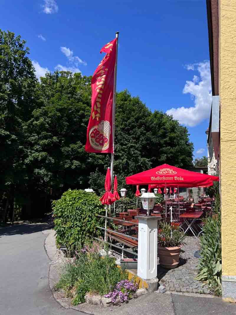 Sonniger Biergarten der Wurz´n in Vilshofen mit roter Fahne, Sonnenschirmen und gemütlichen Holztischen.